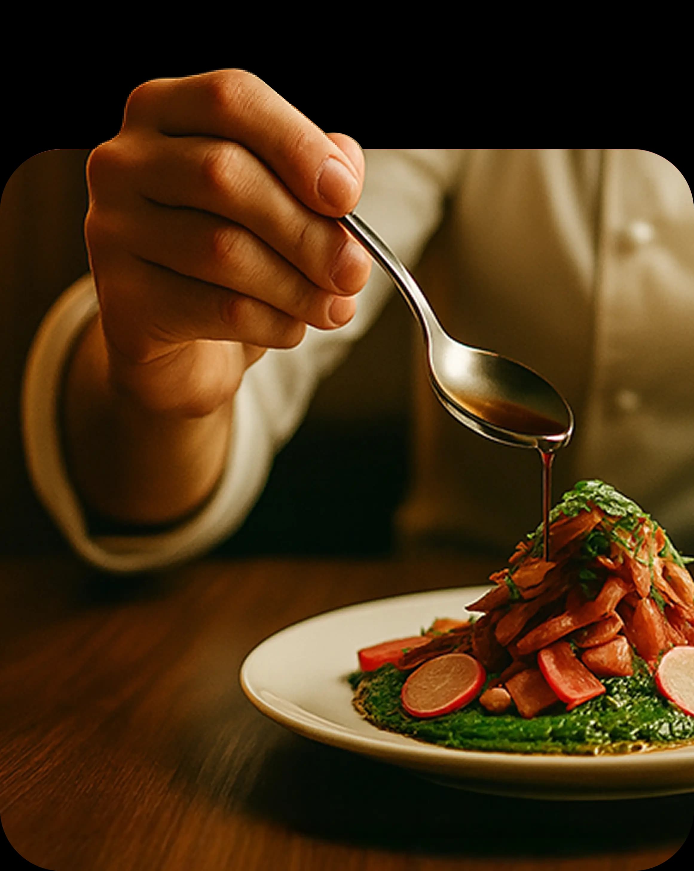 Рука держит ложку, из которой стекает соус на изысканно сервированное блюдо на тарелке, на фоне приглушённого интерьера и человека в белой рубашке.