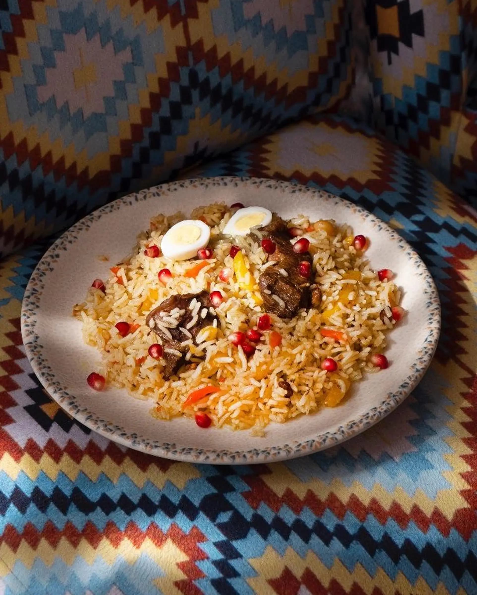 Тарелка плова с кусочками баранины, вареными яйцами, гранатовыми зернами и овощами на узорчатой синей ткани.