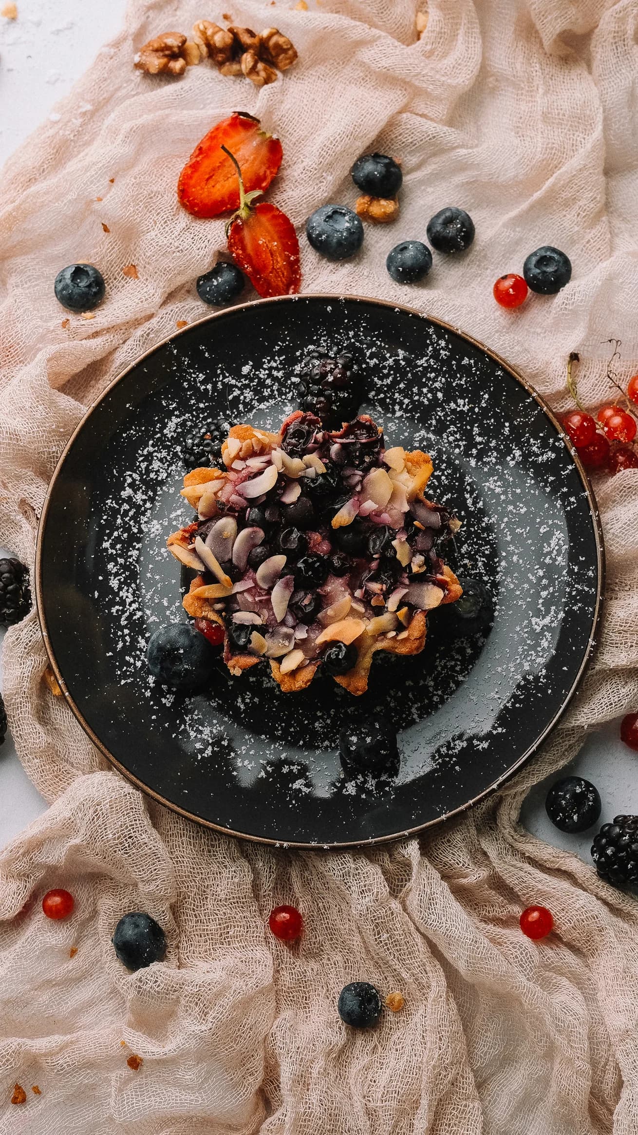 Тарталетка с ягодами, миндальными лепестками и сахарной пудрой на чёрной тарелке.