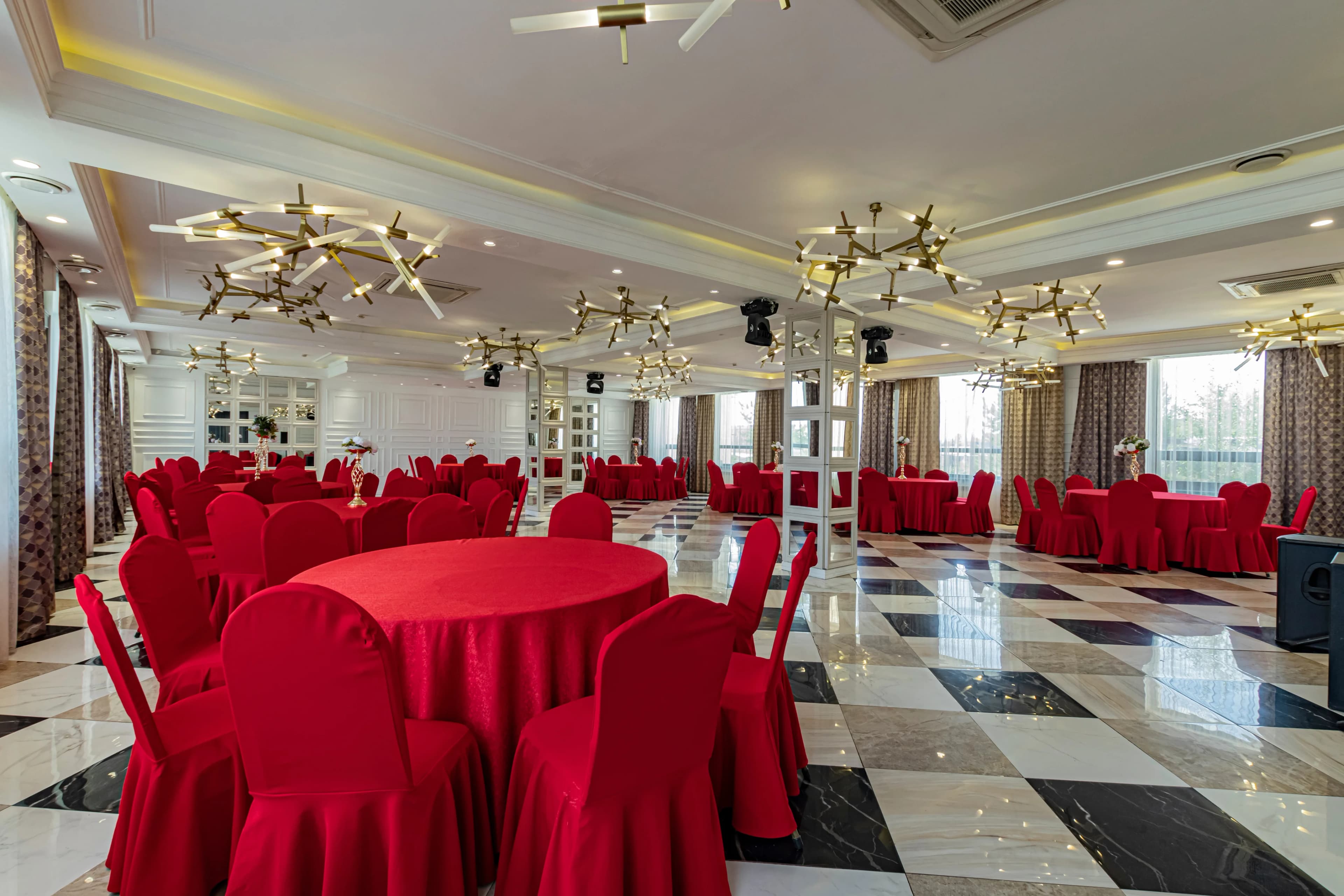Просторный банкетный зал с круглым расположением столов, красными скатертями и современными люстрами.