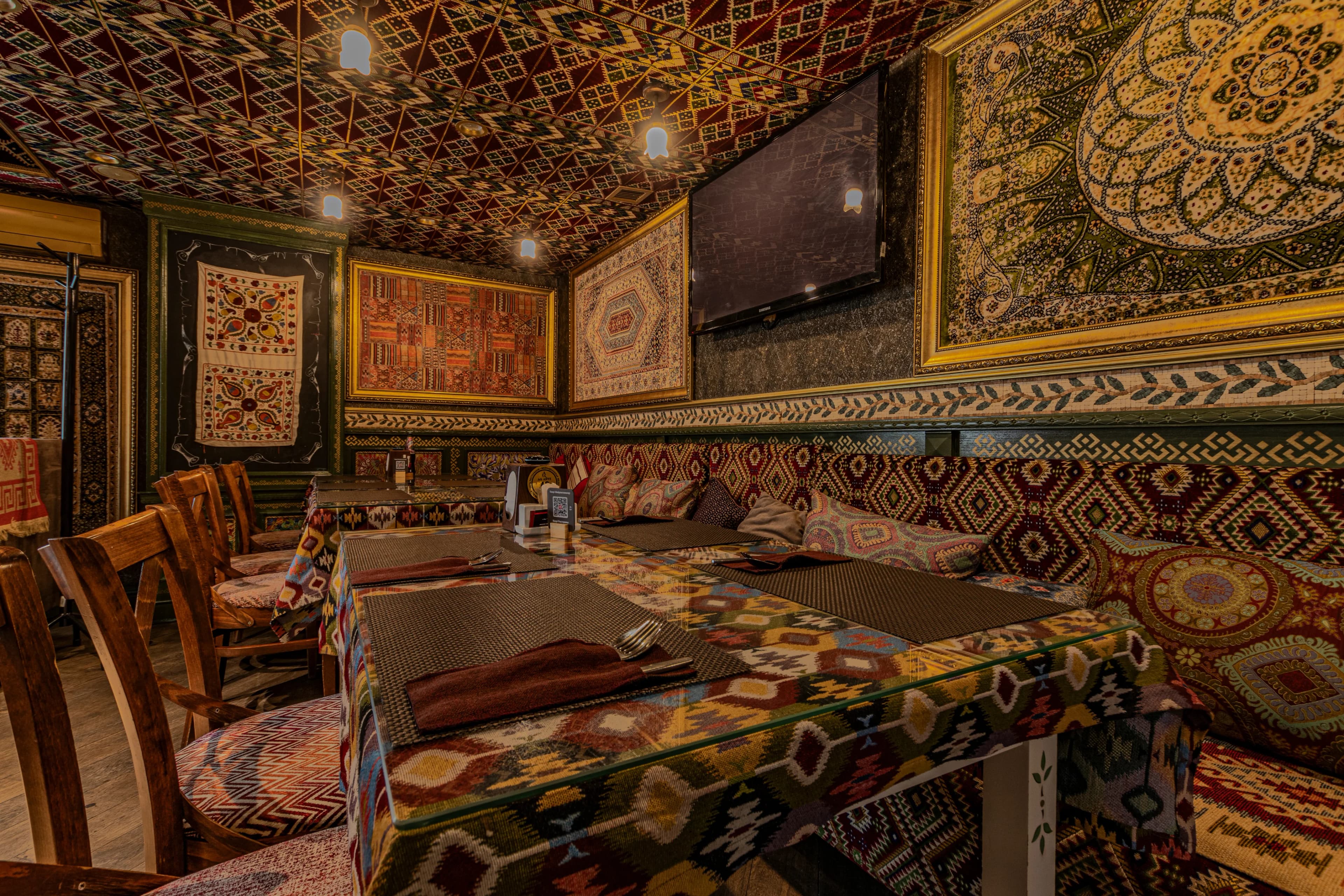 Восточный интерьер ресторана с коврами на стенах и потолке, деревянными стульями и узорными скатертями.