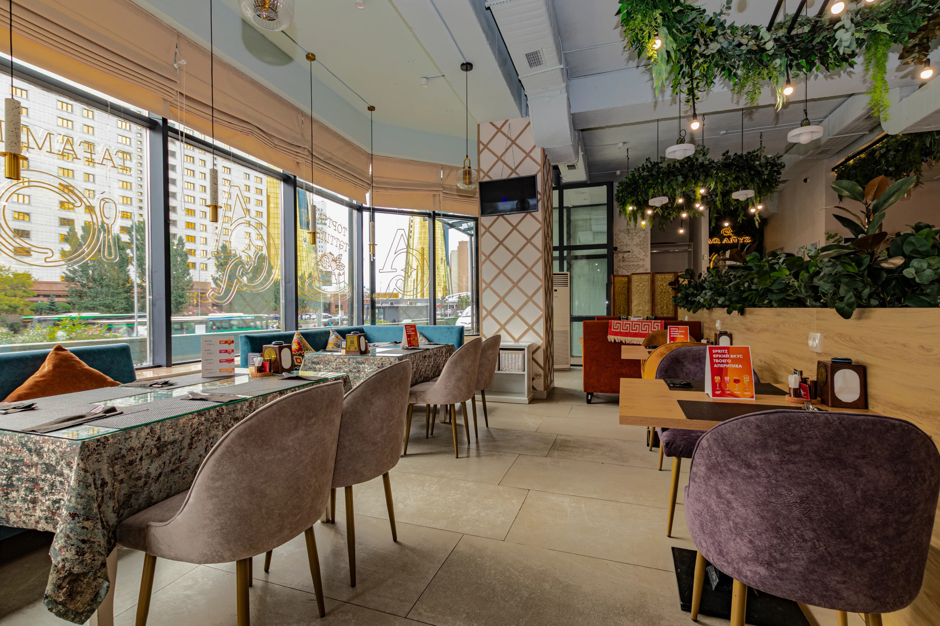Светлый ресторан с панорамными окнами, зелёными растениями и уютными диванами.