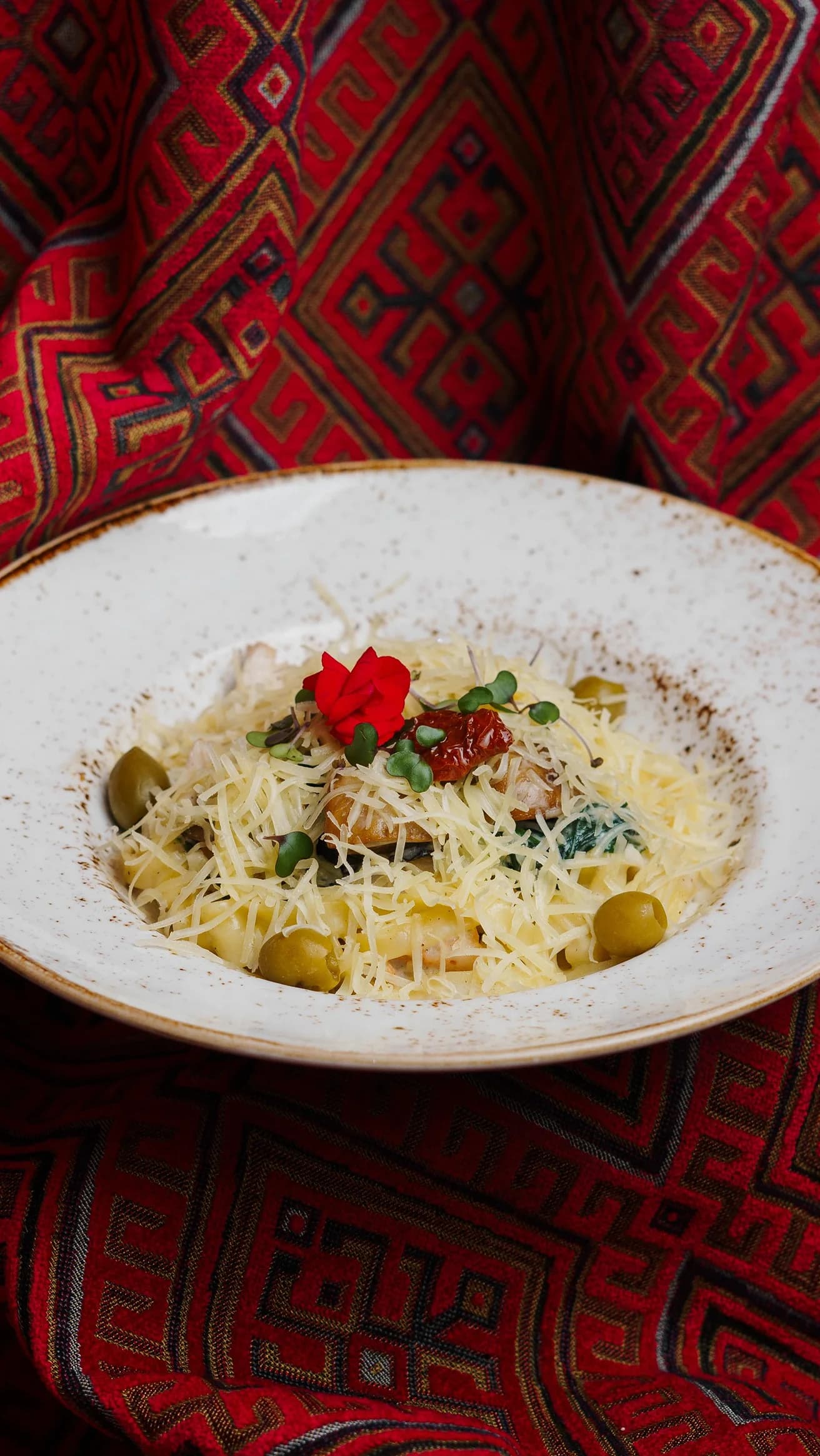 Тарелка пасты с оливками, сыром и овощами, украшенная красным цветком.