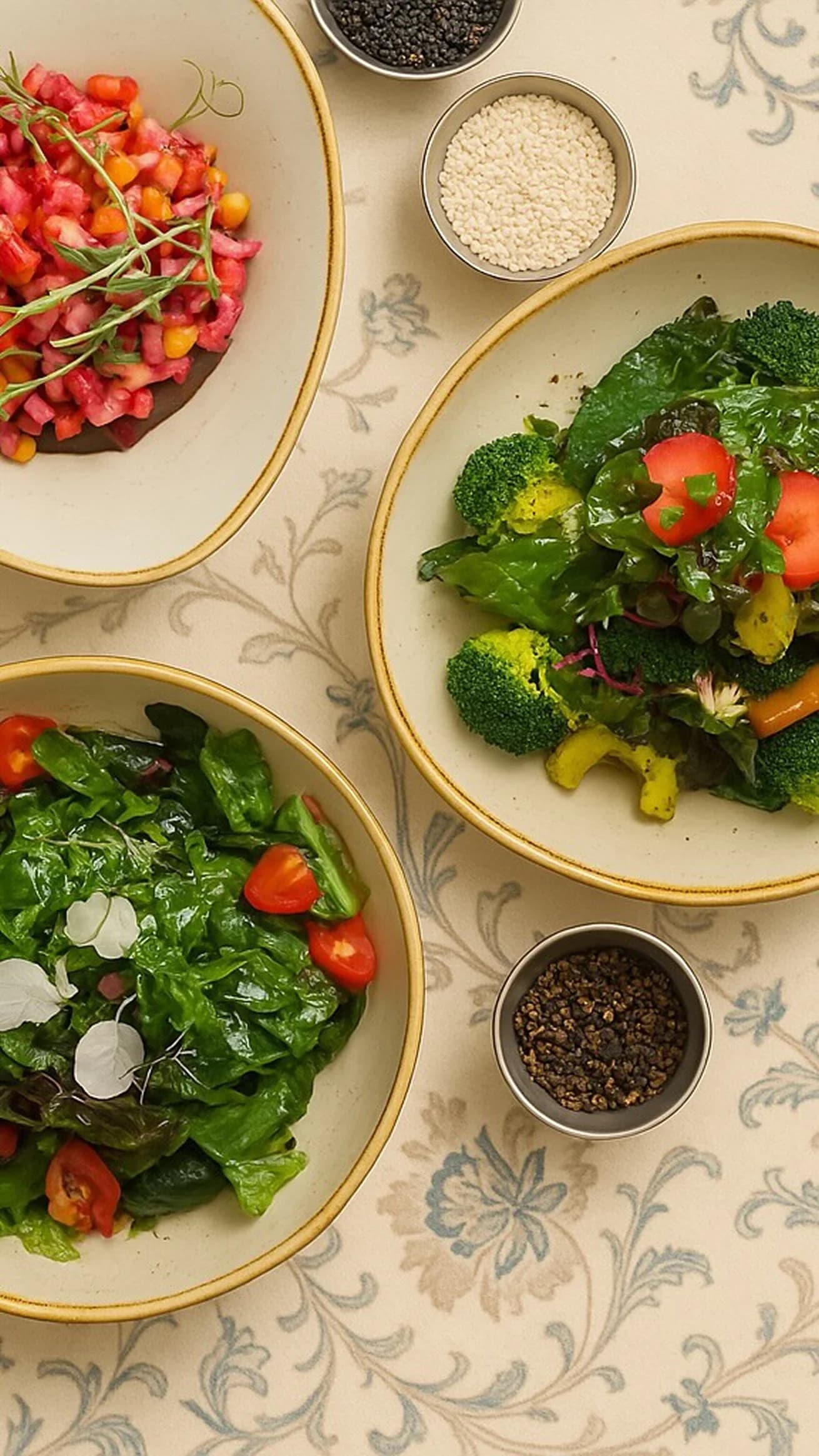 Несколько тарелок с овощными салатами и мисочками специй на светлом столе.