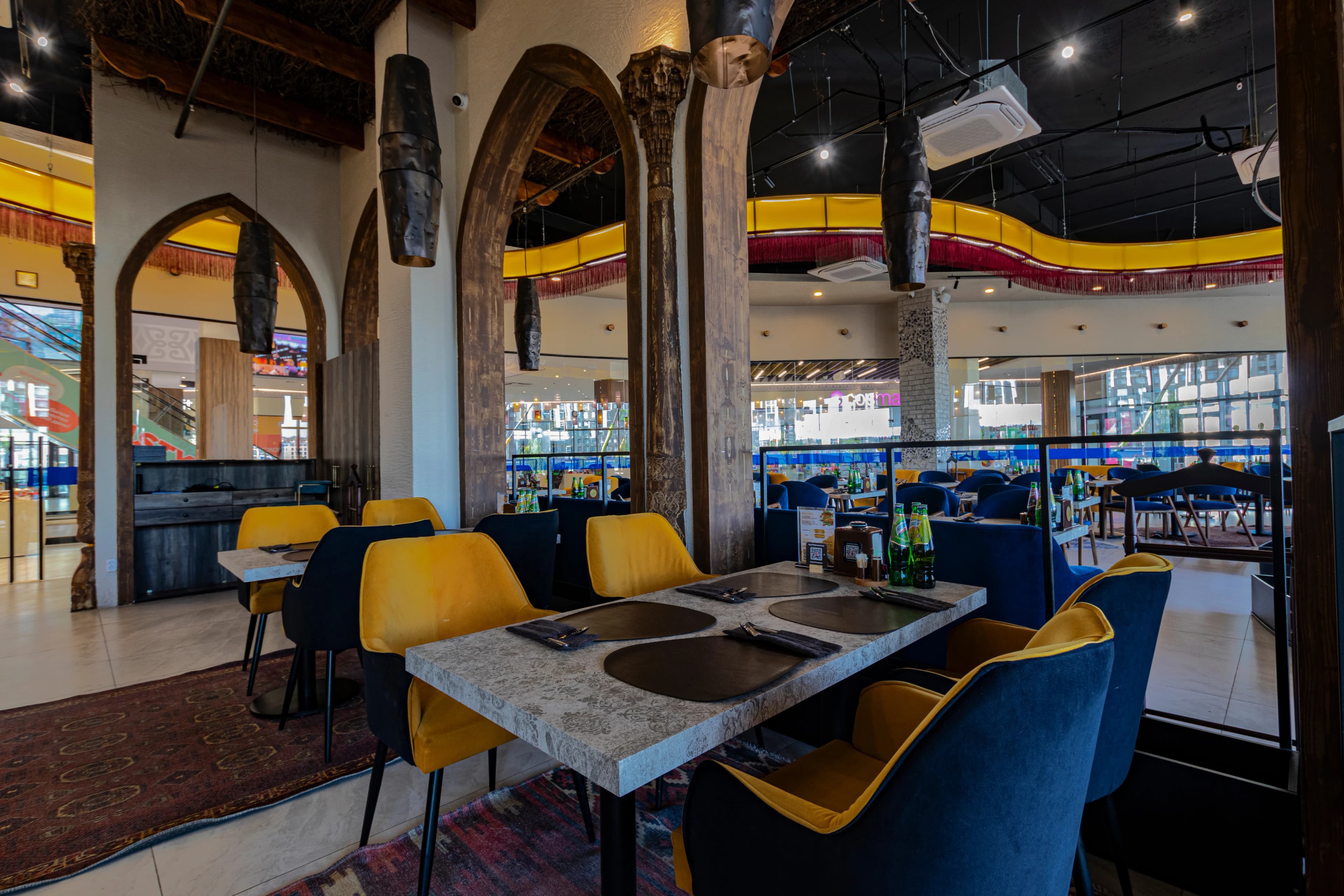 Панорамный вид на пустой просторный зал ресторана с современным интерьером: ряды столов со светлыми столешницами и стульями жёлтого и тёмно‑синего цвета, над ними висят цилиндрические подвесные светильники, на столах расставлены бутылки, бокалы и приборы, потолок украшен извилистыми красно‑жёлтыми конструкциями, а в глубине видны арочные проёмы и барная стойка.